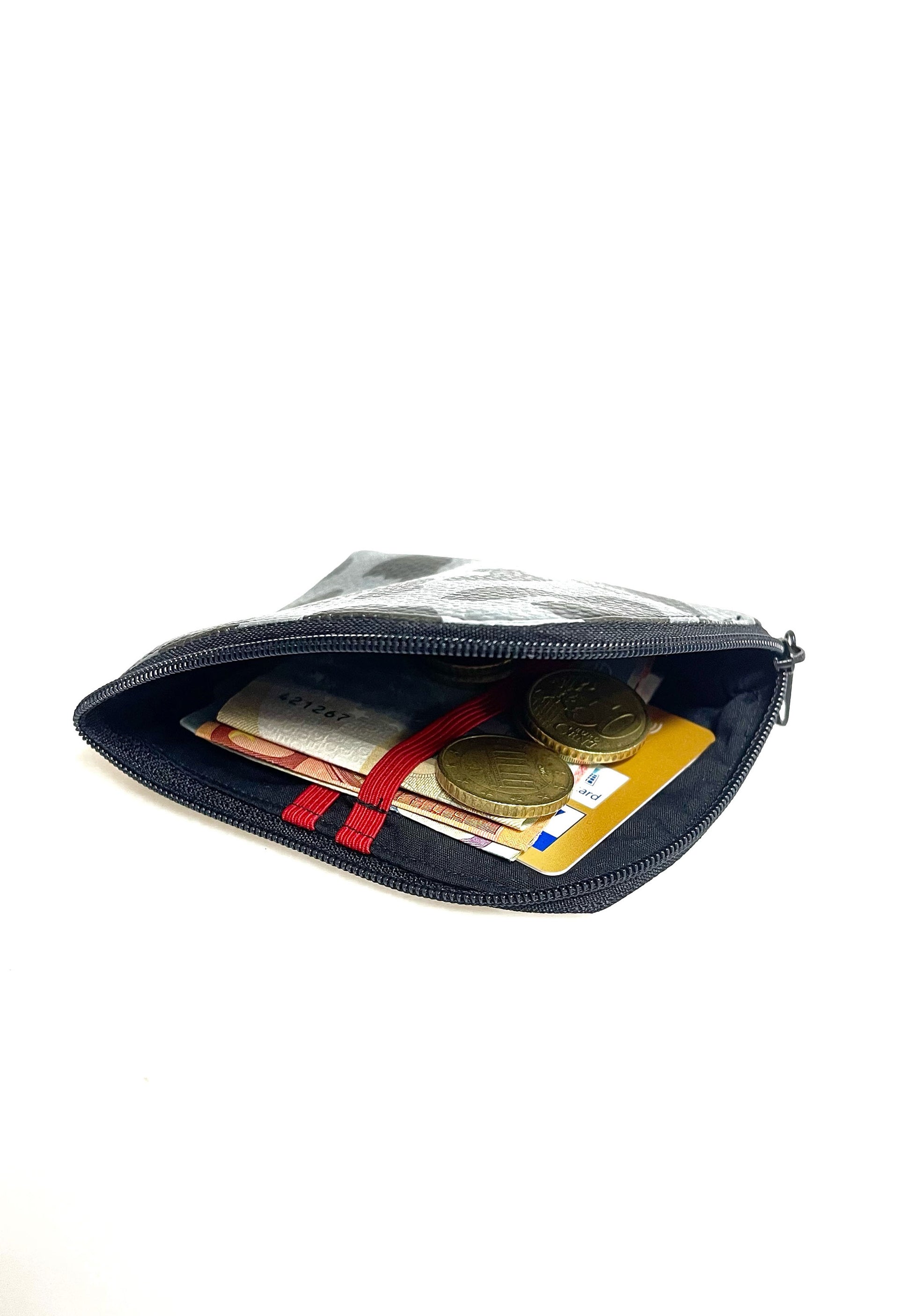 Kleine Geldbörse aus grauem Kunstleder mit schwarzen Leopradenflecken. Sie ist rechteckig mit einer abgerundeten Ecke. An zwei Seiten ist sie mit einem schwarzen Reißverschluss geschlossen. Ansicht mit offenem Reißverschluss. Man sieht das schwarze Futter und die roten Gummibänder.  Außerdem eine EC-Karte und Geldscheine, die in den Gummibändern stecken und Münzen, die darauf liegen. 