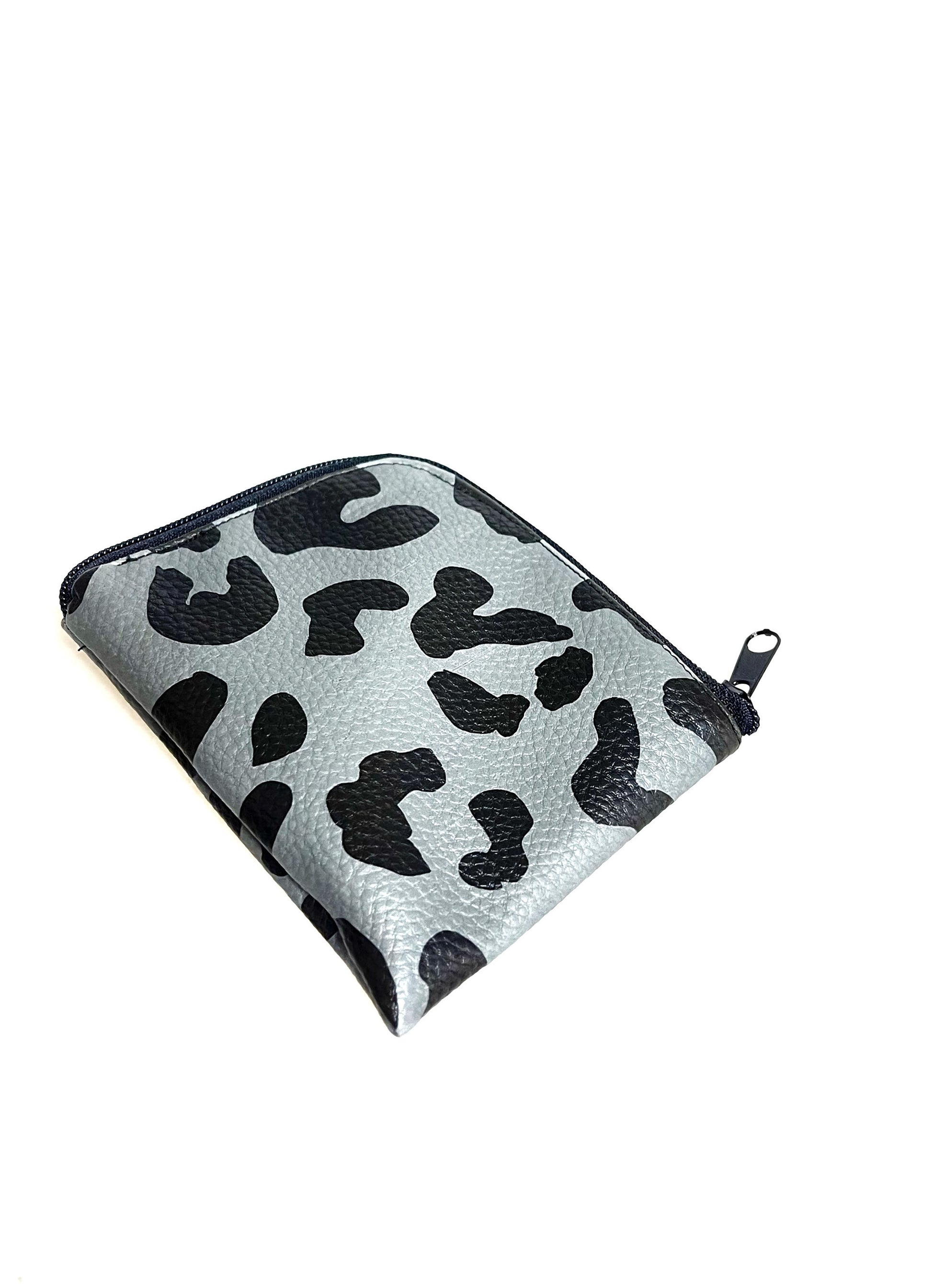 Kleine Geldbörse aus grauem Kunstleder mit schwarzen Leopradenflecken. Sie ist rechteckig mit einer abgerundeten Ecke. An zwei Seiten ist sie mit einem schwarzen Reißverschluss geschlossen. Ansicht von schräg unten. 