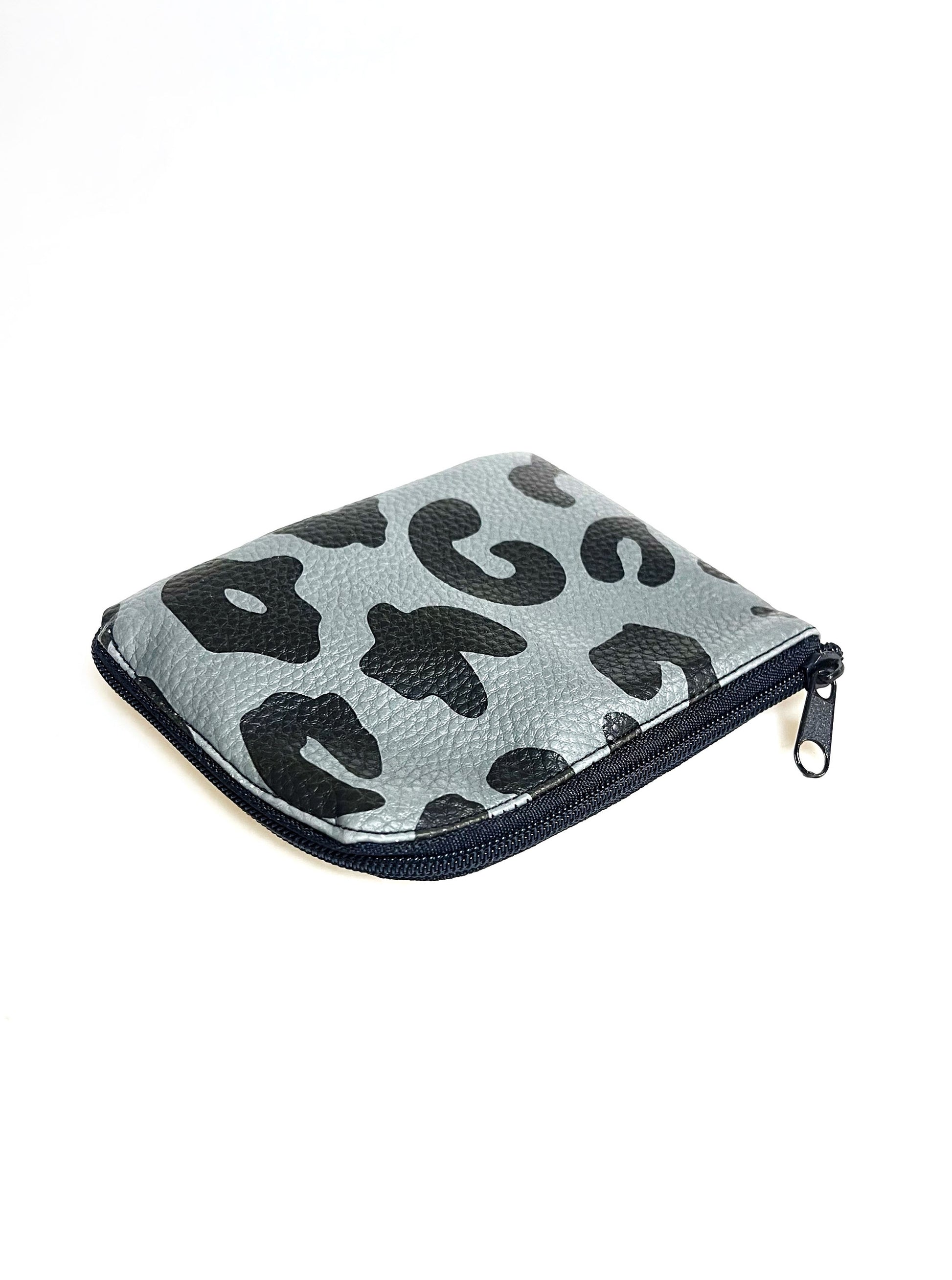 Kleine Geldbörse aus grauem Kunstleder mit schwarzen Leopradenflecken. Sie ist rechteckig mit einer abgerundeten Ecke. An zwei Seiten ist sie mit einem schwarzen Reißverschluss geschlossen. Ansicht von schräg oben. 