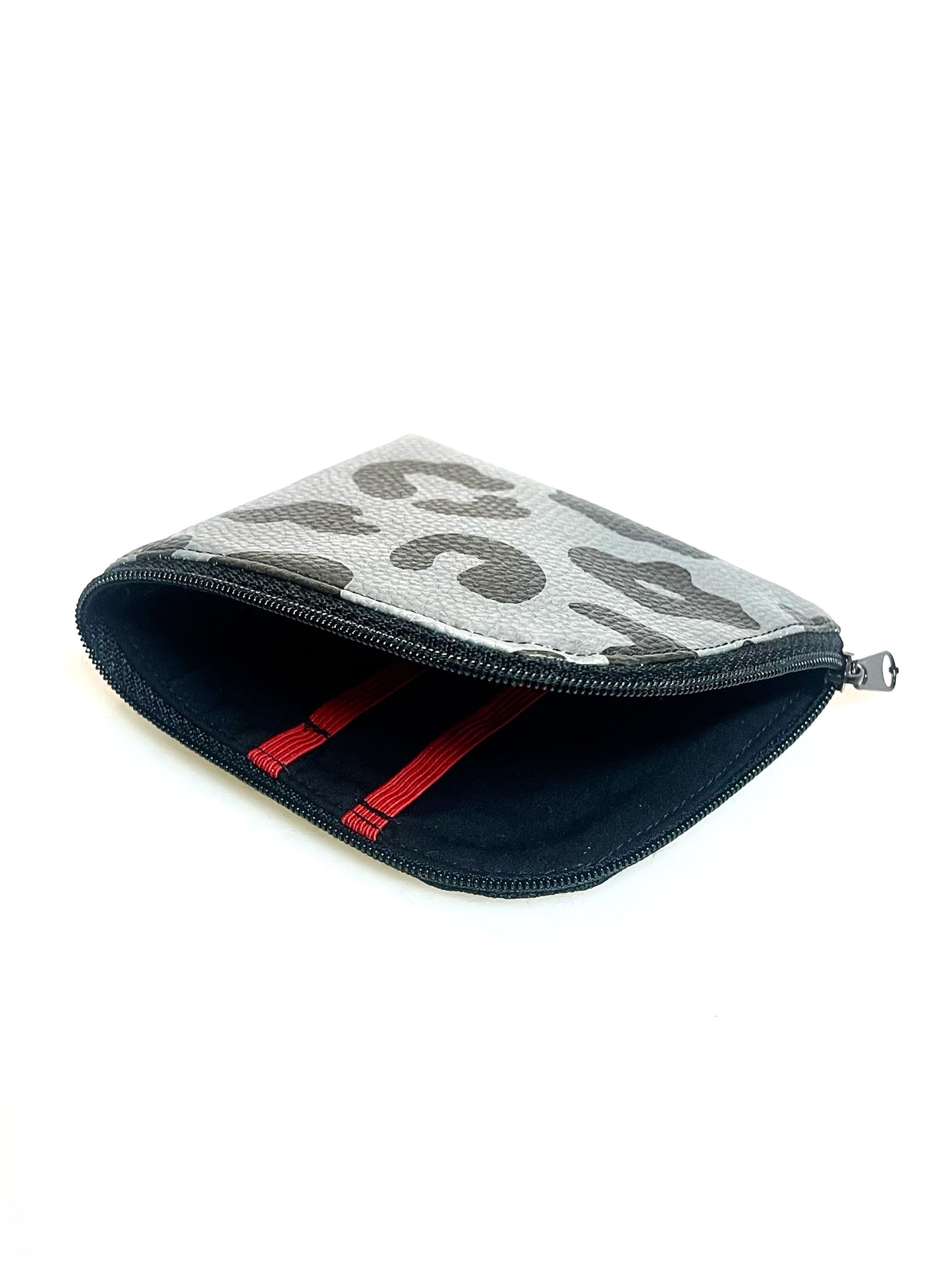 Kleine Geldbörse aus grauem Kunstleder mit schwarzen Leopradenflecken. Sie ist rechteckig mit einer abgerundeten Ecke. An zwei Seiten ist sie mit einem schwarzen Reißverschluss geschlossen. Ansicht mit offenem Reißverschluss. Man sieht das schwarze Futter und die roten Gummibänder.  