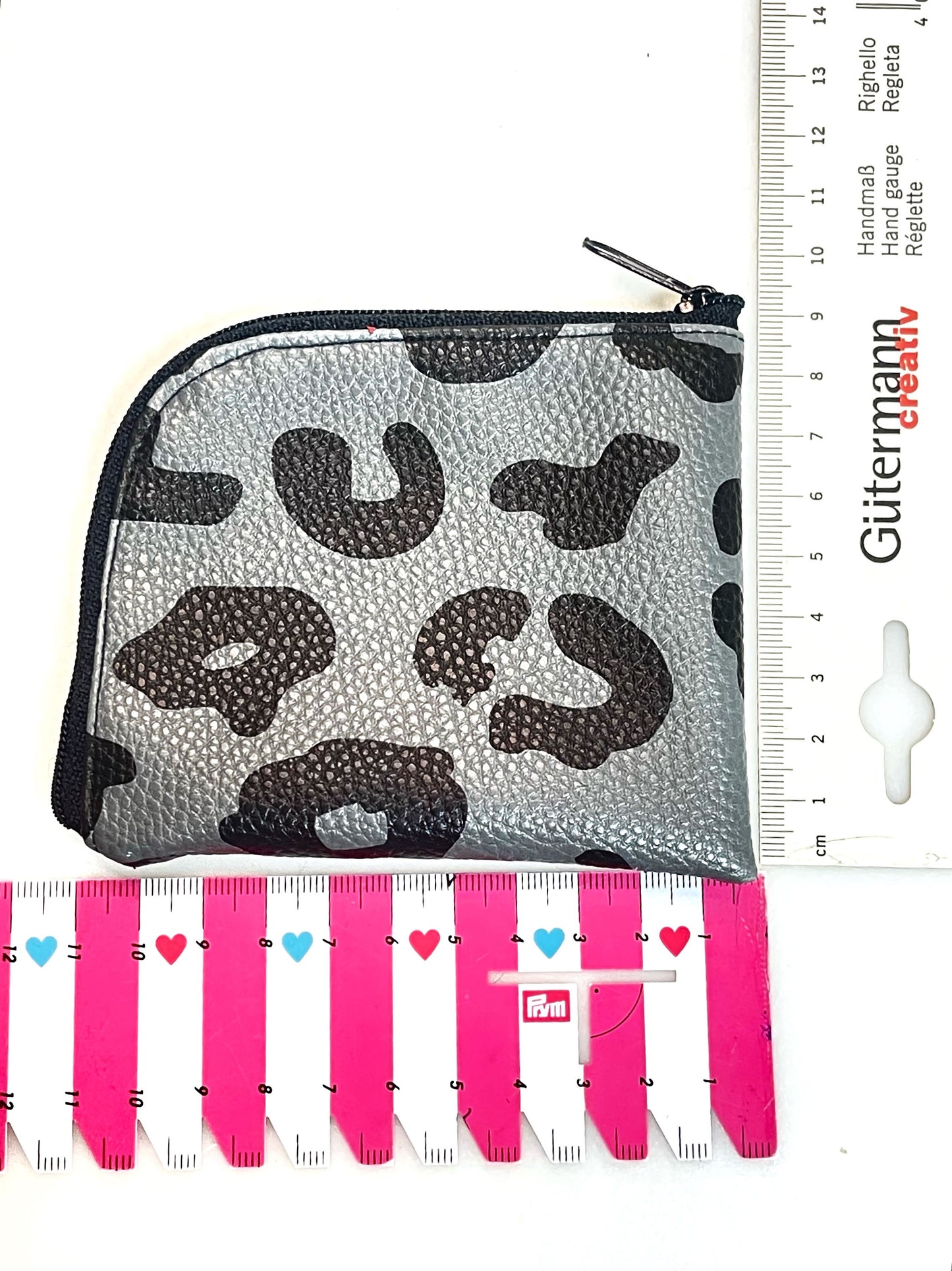 Kleine Geldbörse aus grauem Kunstleder mit schwarzen Leopradenflecken. Sie ist rechteckig mit einer abgerundeten Ecke. An zwei Seiten ist sie mit einem schwarzen Reißverschluss geschlossen. Draufsicht mit 2 Linealen unten und an der Seite, um die Größe besser zu erkennen. 