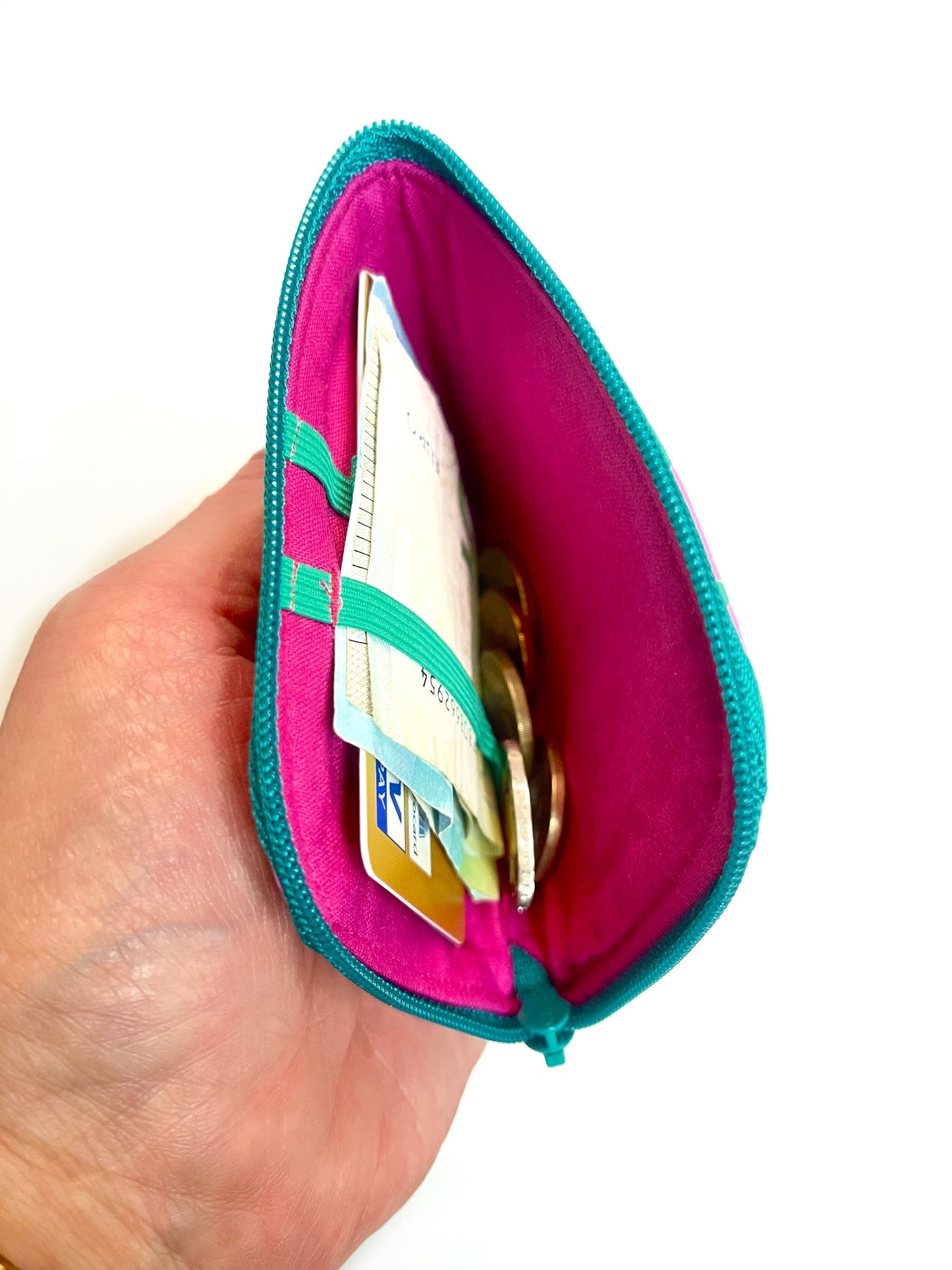 Eine Hand hält die geöffente Börse. Man schaut auf das pinke Innenfutter und die grünen Gummbänder. In den Bändern stecken Geldscheine und eine EC-Karte. Kleingeld liegt daneben.  