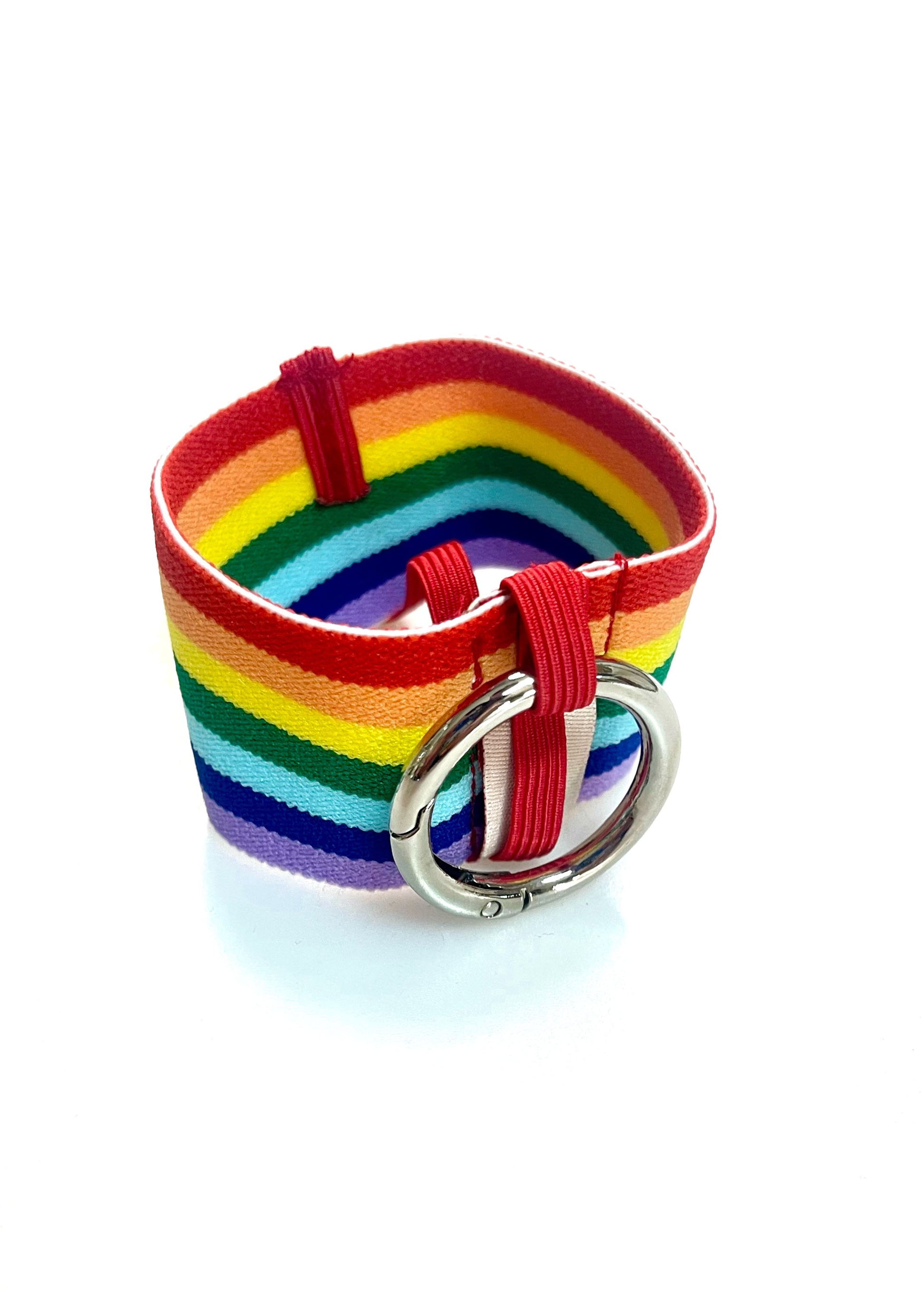 Ein Becherhalter aus einem breiten Gummiband mit Streifenmuster in Regenbogenfarben. Er hat einem runden, silbernen Karabinerhaken. Das Gummiband ist zu einem Kreis zusammengenäht. Der Karabiner ist an einem schmalen Gummiband befestigt, das unten um das breite Gummiband herum geht. rnen