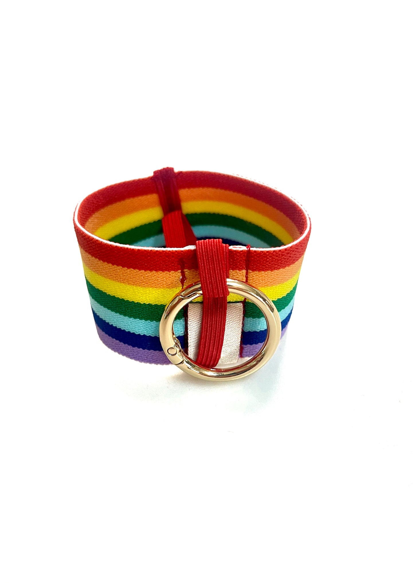 Ein Becherhalter aus einem breiten Gummiband mit Streifenmuster in Regenbogenfarben. Er hat einem runden, goldenen Karabinerhaken. Das Gummiband ist zu einem Kreis zusammengenäht. Der Karabiner ist an einem schmalen Gummiband befestigt, das unten um das breite Gummiband herum geht. 