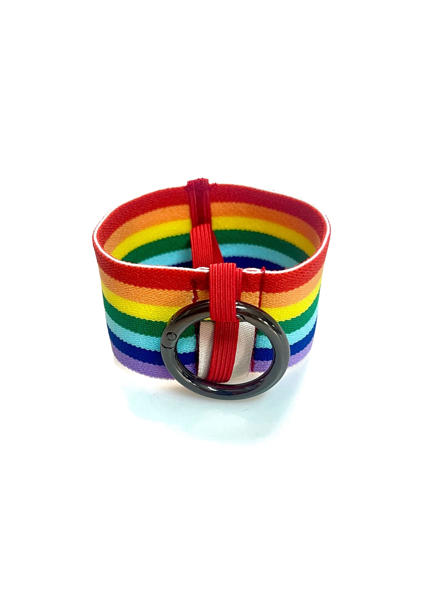 Ein Becherhalter aus einem breiten Gummiband mit Streifenmuster in Regenbogenfarben. Er hat einem runden, gunmetal farbenen Karabinerhaken. Das Gummiband ist zu einem Kreis zusammengenäht. Der Karabiner ist an einem schmalen Gummiband befestigt, das unten um das breite Gummiband herum geht. 