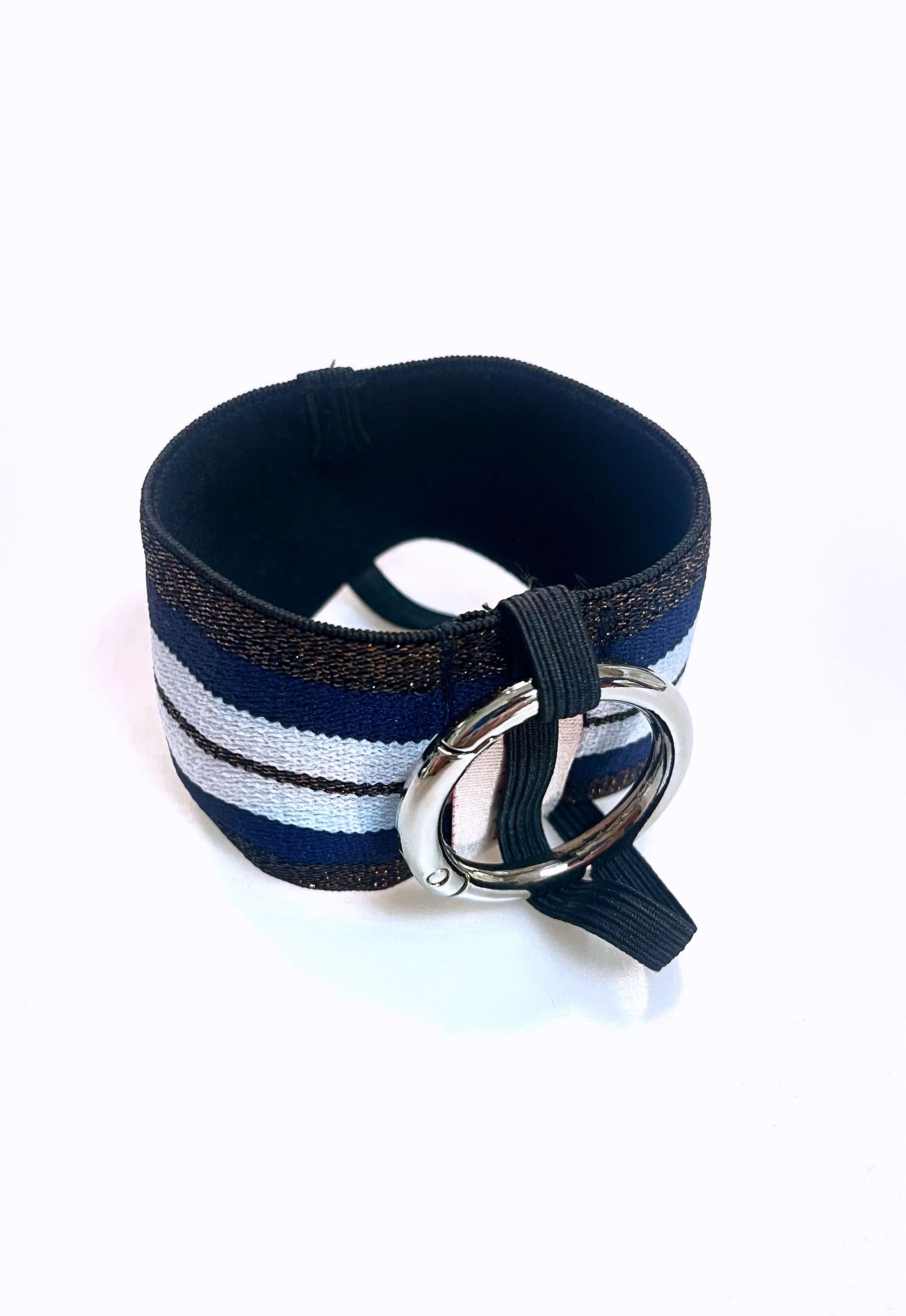 Ein Becherhalter aus einem breiten Gummiband mit Streifenmuster in weiß, dunkelblau und kupfer mit schwarz-Glitzer. Er hat einem runden, silbernen Karabinerhaken. Das Gummiband ist zu einem Kreis zusammengenäht. Der Karabiner ist an einem schmalen Gummiband befestigt, das unten um das breite Gummiband herum geht. 