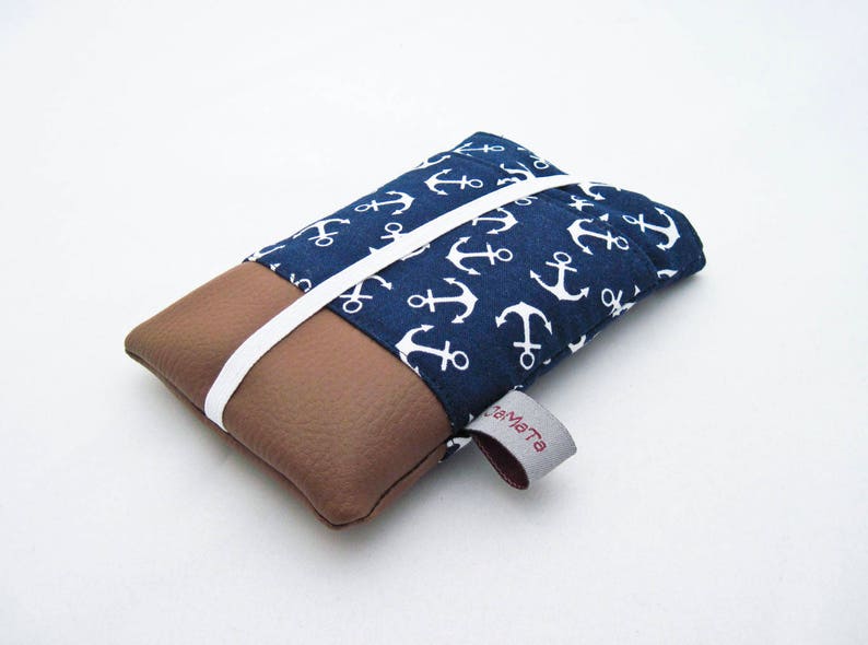 Eine Handytasche aus dunkelblauer Baumwolle mit weißen Ankern oben und braunem Kunstleder unten. Die Tasche hat eine aufgesetzte Tasche aus dem Ankerstoff und ist mit weißem Gummiband geschlossen. 
Aufnahme seitlich von unten 