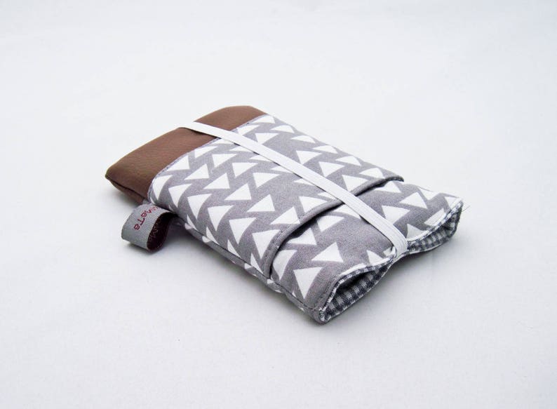 Eine Handytasche aus grauer Baumwolle mit weißen Dreiecken oben und braunem Kunstleder unten. Die Tasche hat eine aufgesetzte Tasche aus dem Stoff mit den Dreiecken und ist mit weißem Gummiband geschlossen. 
Aufnahme seitlich von oben.