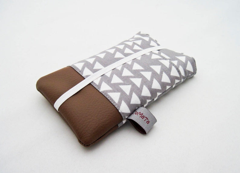 Eine Handytasche aus grauer Baumwolle mit weißen Dreiecken oben und braunem Kunstleder unten. Die Tasche hat eine aufgesetzte Tasche aus dem Stoff mit den Dreiecken und ist mit weißem Gummiband geschlossen. 
Aufnahme seitlich von unten.