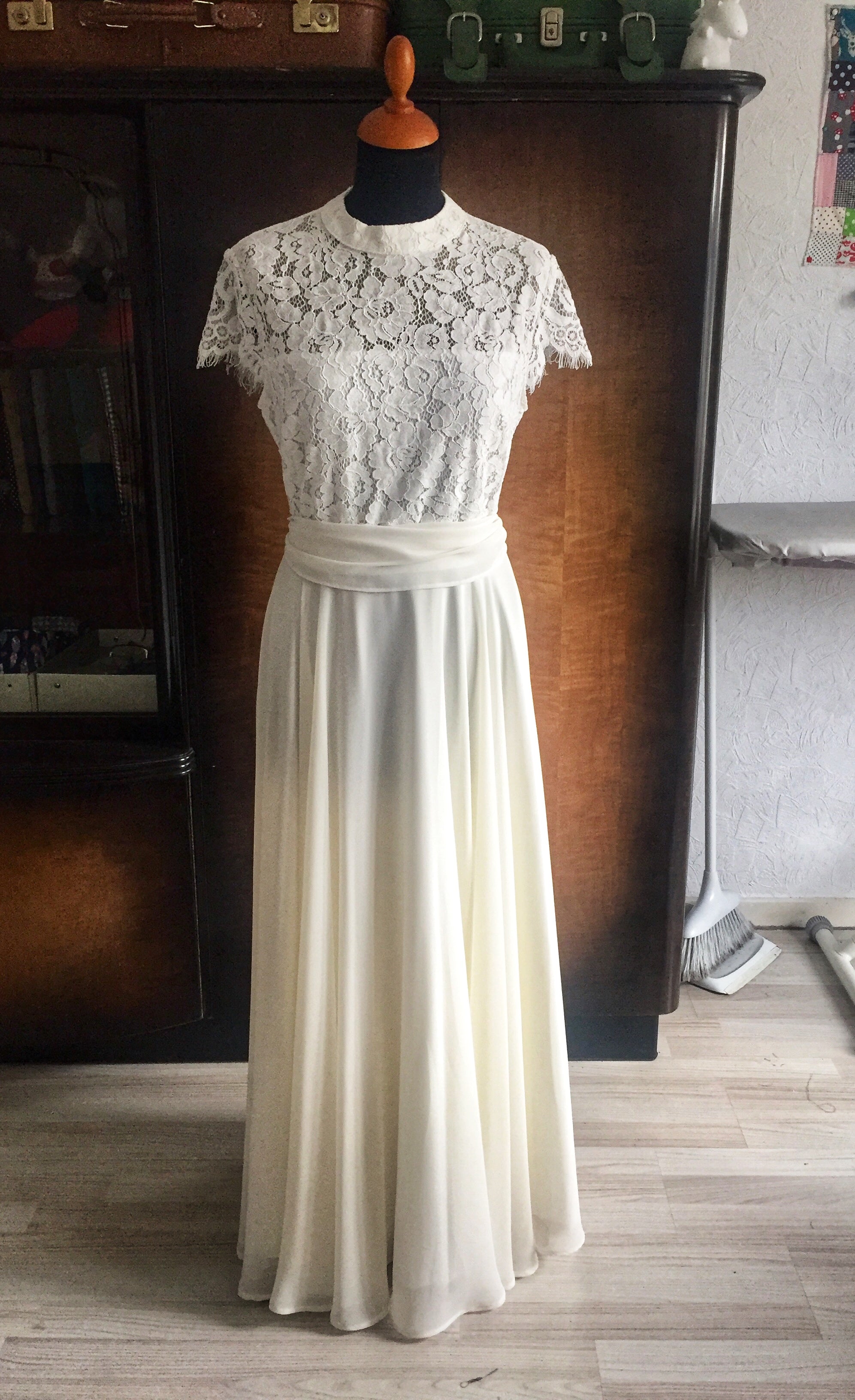 Brautkleid mit einem kurzärmligen Oberteil aus weißer Spitze, das bis über die Brust abgefütter ist. Der Rock ist lang und fließend aus mehreren Chiffonlagen. Das Kleid hat eine Schärpe aus dem Chiffonstoff. Es ist auf einer Schneiderpuppe fotografiert, die vor einem dunklen Vintagschrank steht, mit Vintage Koffern darauf.