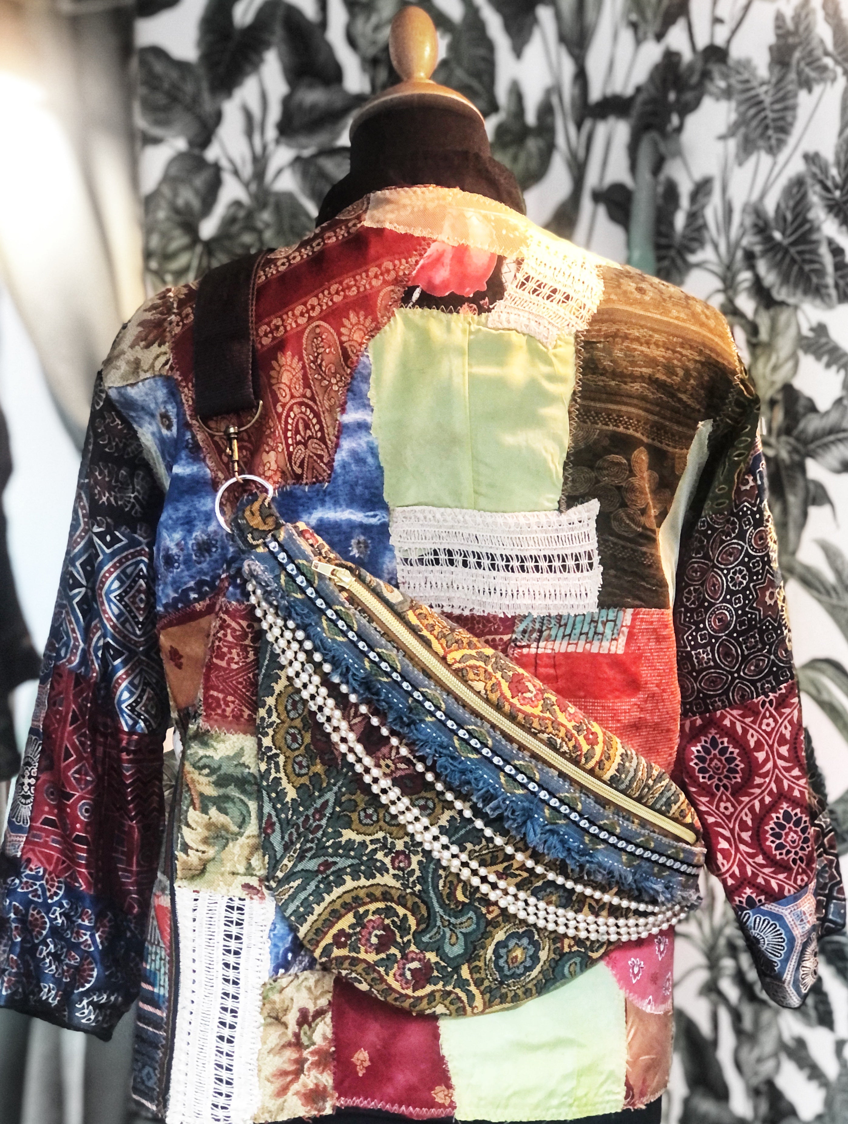 Rückansicht einer Jacke auf einer Schneiderpuppe, die aus verschiedenen Stoffresten zusammengesetzt ist. Über der Jacke hängt eine Bauchtasche, die ebenfalls aus Stoffresten, Borten und Ketten gestaltet ist.