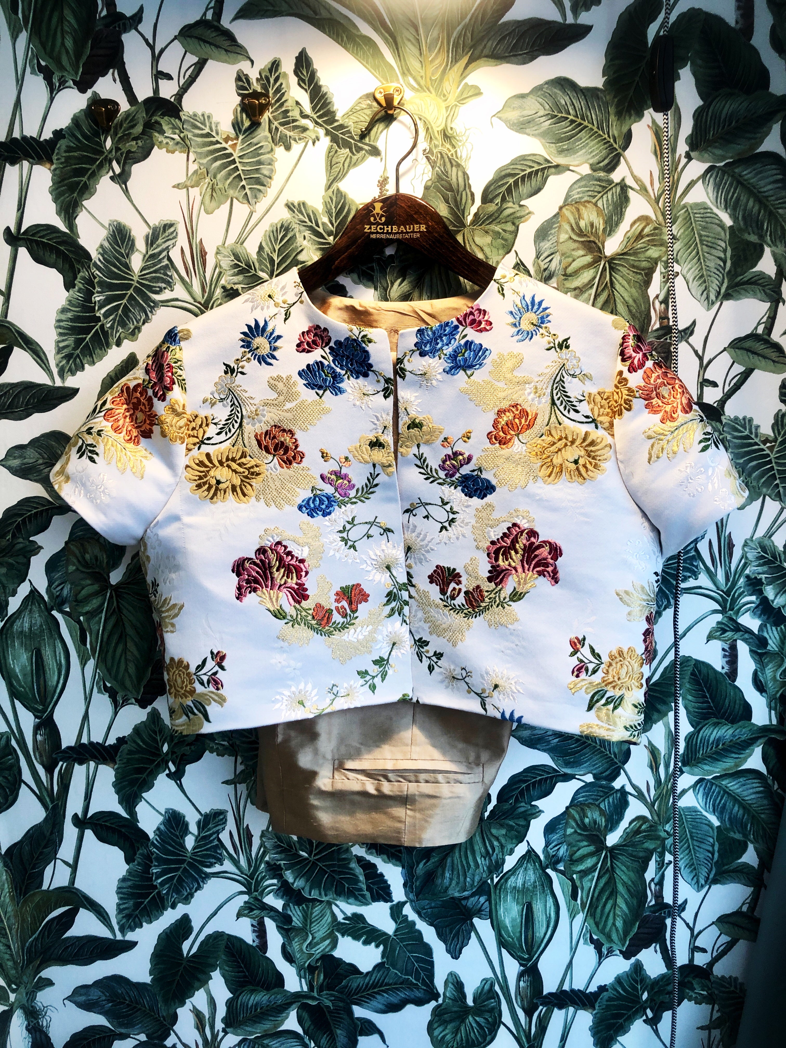 Eine kurze Bolerojacke mit kurzen Ärmlen auf einem Bügel. Die Jacke ist weiß und mit bunten und goldenen Blumen bestickt. Unter der Jacke schaut eine goldene Hose hervor. Beides hängt vor einer Wand mit grüner Blättertapete.