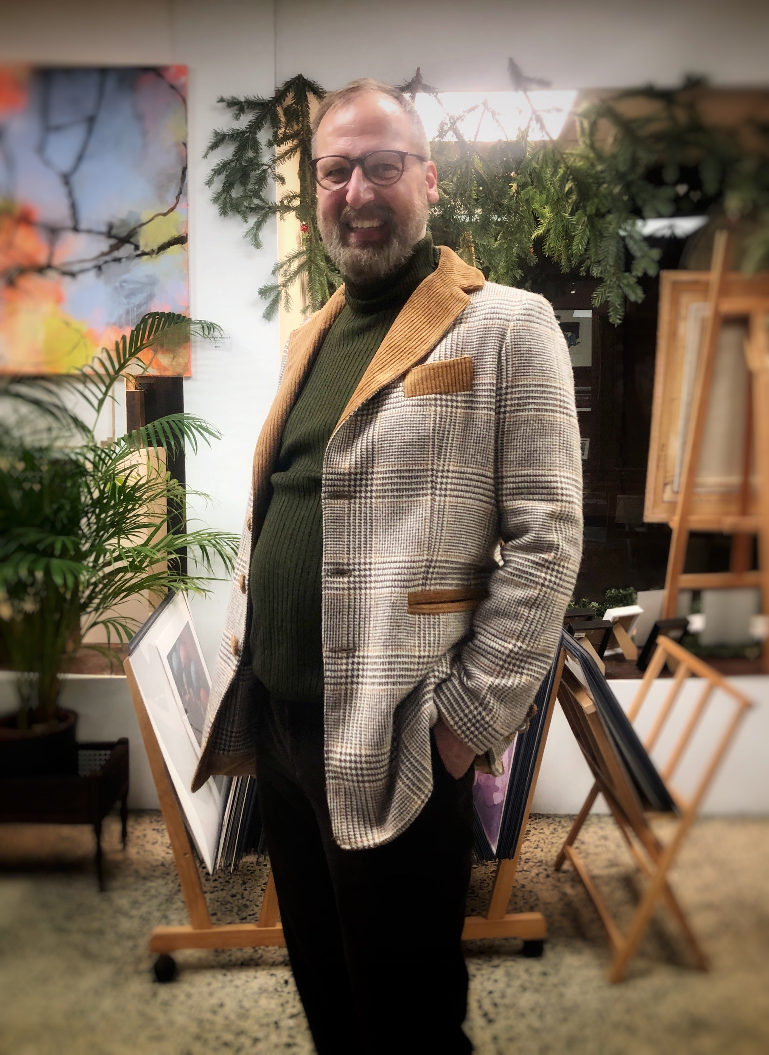 Ein Mann in einem Atelier mit Bildern und Pflanzen. Der Mann hat einen Bart und eine Brille. Er lacht in die Kamera. Er trägt ein beiges Jacket mit braunen Karos und einem Revers und Taschen aus braunem Breitcord über einem dunkelgrünen Rollkragenpullover zu einer dunklen Hose.