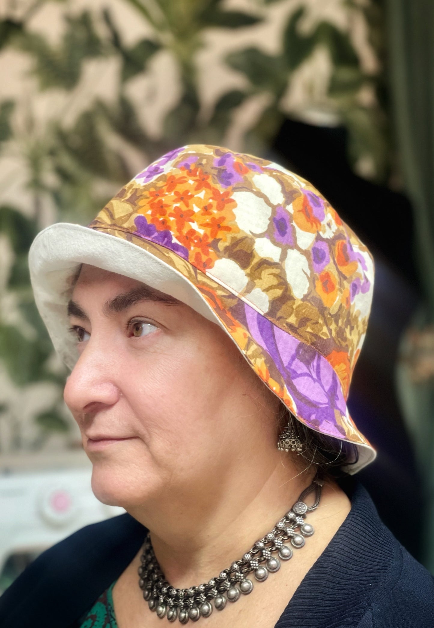 Portrait einer Frau mit brauen Augen und dunklen Augenbrauen im Profil. Sie trägt einen Fischerhut, den sie im Nähkurs selbst genäht hat. Der Hut ist außen aus einem Vintage-Stoff mit großen Blumen in senfgelb, braub, lila, orange und creme und innen aus cremeweißer Baumwolle. 
Außerdem trägt sie indischen Silberschmuck und eine schwarze Strickjacke. Sie sitzt vor einer Wand mit grüner Blättertapete. Der Hintergrund ist unscharf. 