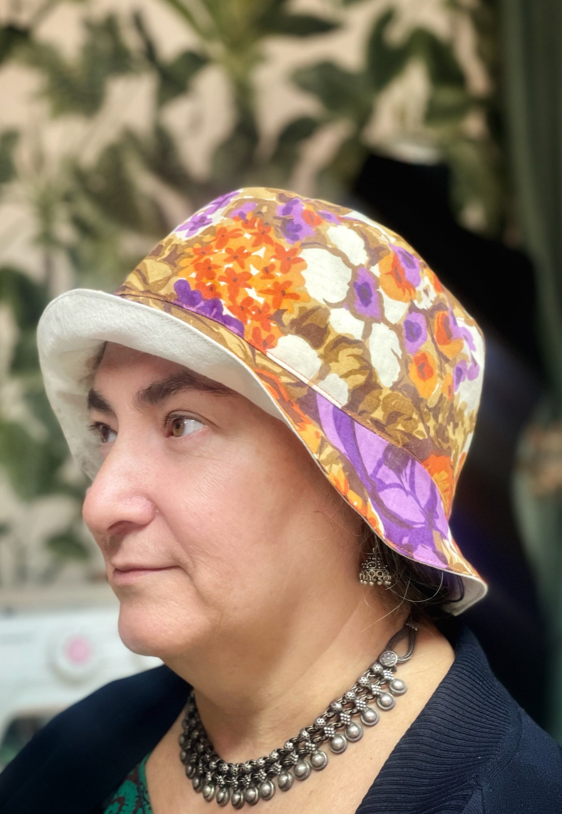 Portrait einer Frau mit brauen Augen und dunklen Augenbrauen im Profil. Sie trägt einen Fischerhut, den sie im Nähkurs selbst genäht hat. Der Hut ist außen aus einem Vintage-Stoff mit großen Blumen in senfgelb, braub, lila, orange und creme und innen aus cremeweißer Baumwolle. 
Außerdem trägt sie indischen Silberschmuck und eine schwarze Strickjacke. Sie sitzt vor einer Wand mit grüner Blättertapete. Der Hintergrund ist unscharf. 