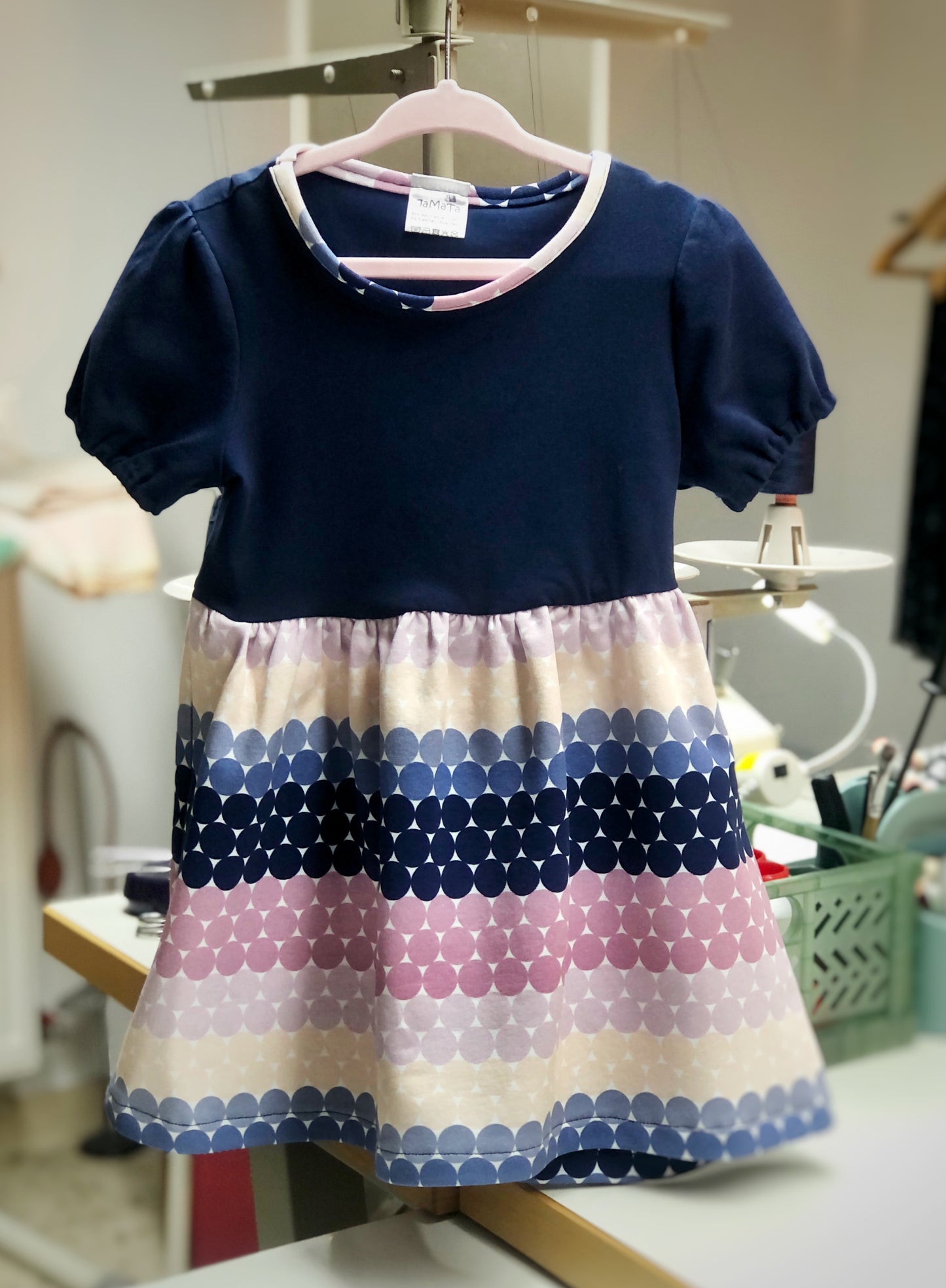 Ein Kinderkleid mit kurzen Puffärmeln. Das Oberteil ist dunkelblau, der Rock aus einem gestreiften Stoff in den Farben, dunkelblau, jeansblau, altrosa, nude und gelb. Dien Streifen haben Farbverläufe und sind in sich gepunktet. Der Rock ist ans Oberteil gekräuselt. 