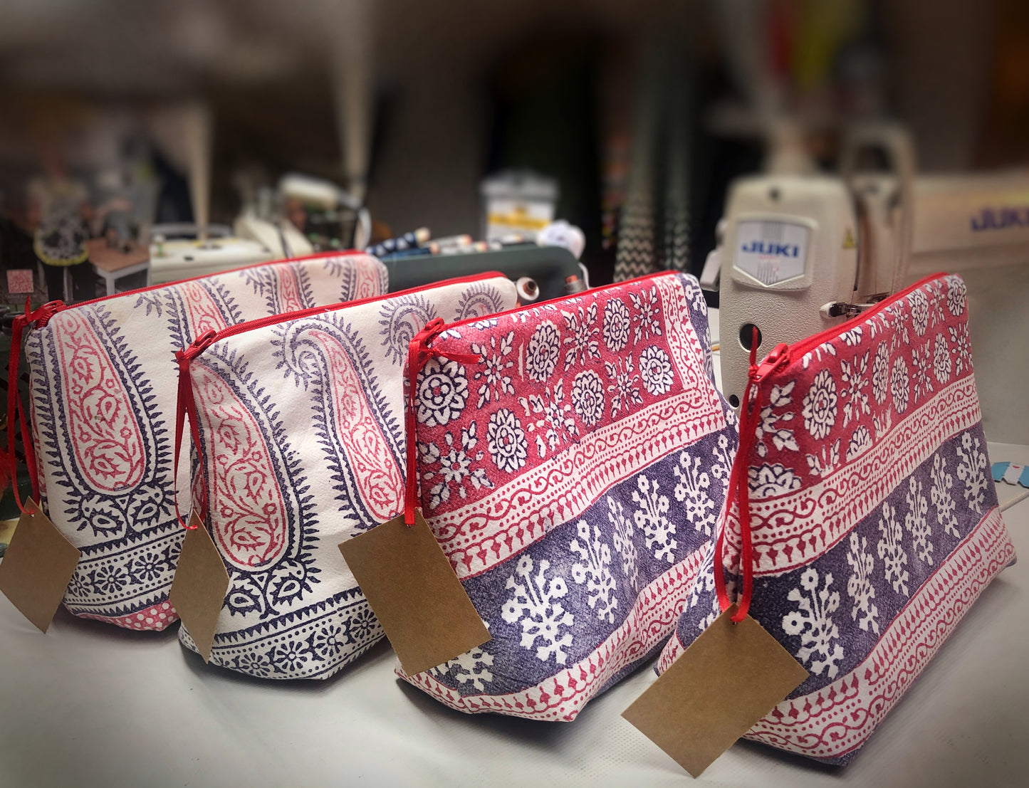 Vier Kosmetiktaschen. die vor einer Nähmaschine stehen. Sie haben ein organisches Muster und sind aus alten Stoffen gemacht. Die Fraben sind naturweiß, blau und rot. An den Reißverschlüssen hängen Etiketten aus waschbarem Papier. 
