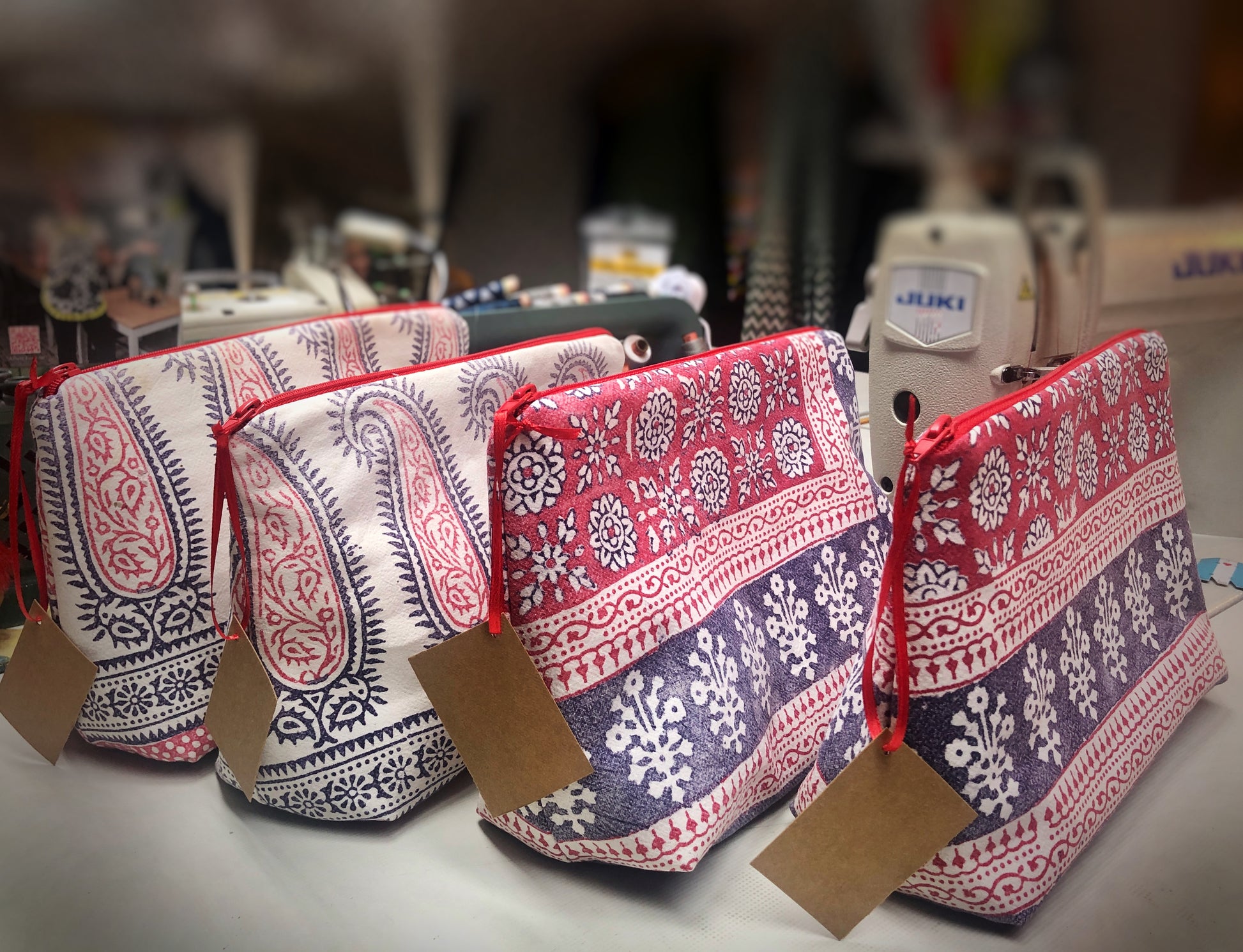 Vier Kosmetiktaschen. die vor einer Nähmaschine stehen. Sie haben ein organisches Muster und sind aus alten Stoffen gemacht. Die Fraben sind naturweiß, blau und rot. An den Reißverschlüssen hängen Etiketten aus waschbarem Papier. 