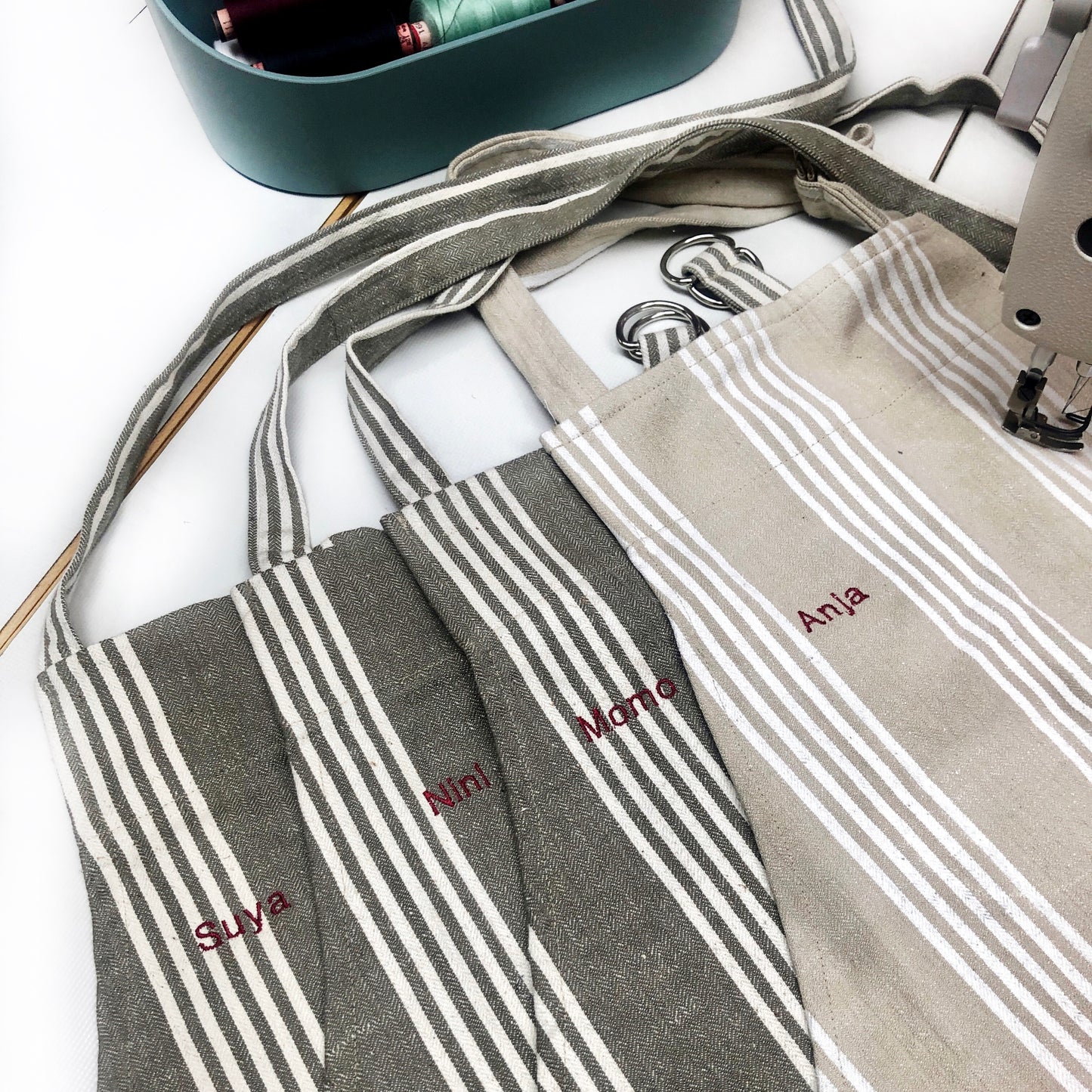 Der obere Teil von vier Schürzen auf einem Nähtisch mit gestickten Namen. Die Stffe sind beige und grau mitz weißen Streifen. Eine Schürze klemmt unter einer Nähmaschine. 