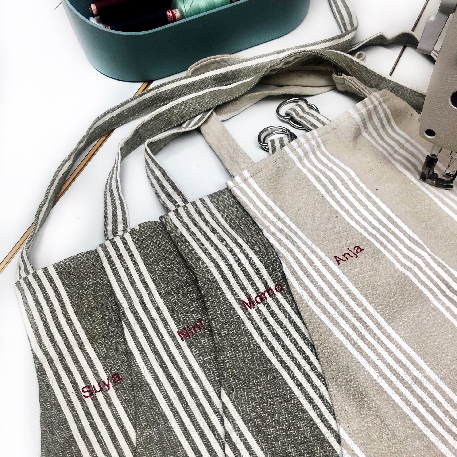 Der obere Teil von vier Schürzen auf einem Nähtisch mit gestickten Namen. Die Stffe sind beige und grau mitz weißen Streifen. Eine Schürze klemmt unter einer Nähmaschine. 