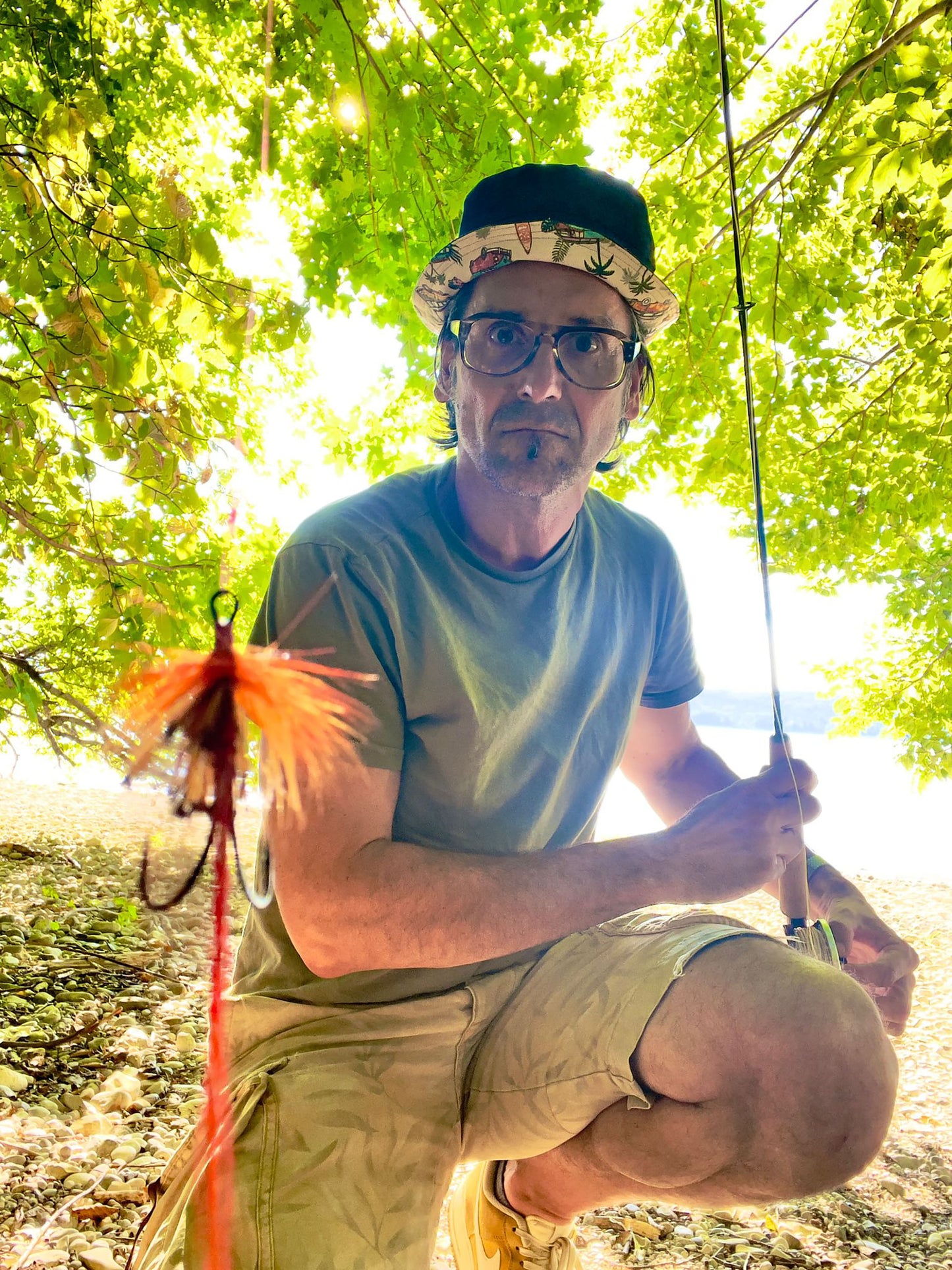 Ein Mann mit Brille, T-Shirt und kurzen Hosen kniet an einem steinigen Seeufer unter einem Baum. Er rägt den Fischerhut mit der Jeansseite außen. Die Krempe des Hutes ist hoch geschlagen, so dass man das Muster mit den Bullis auf der Innenseite sieht. In der Hand hält er eine Angel. 